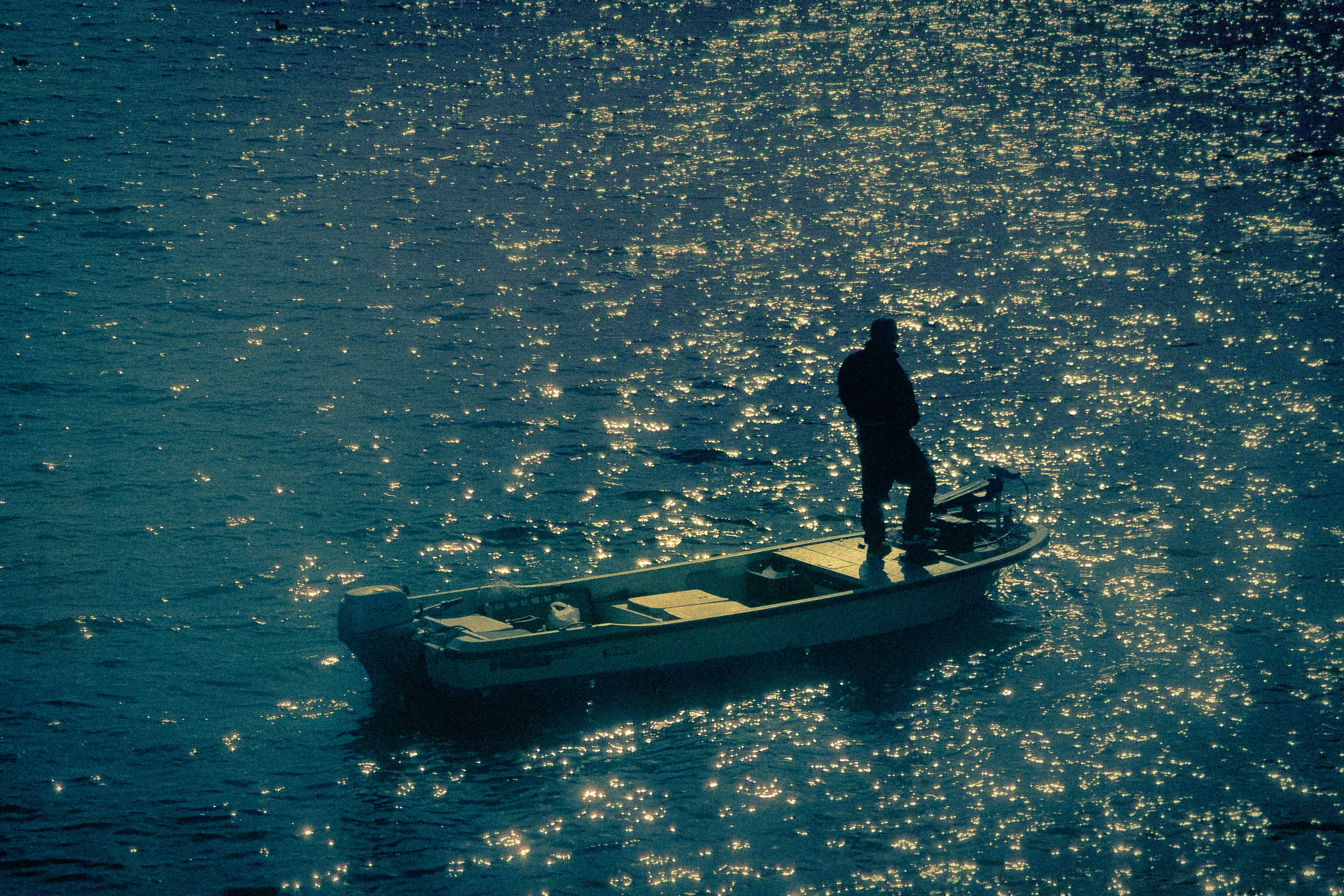 A silhouetted person standing at the bow of a small motorboat on dark water covered in glittering light.